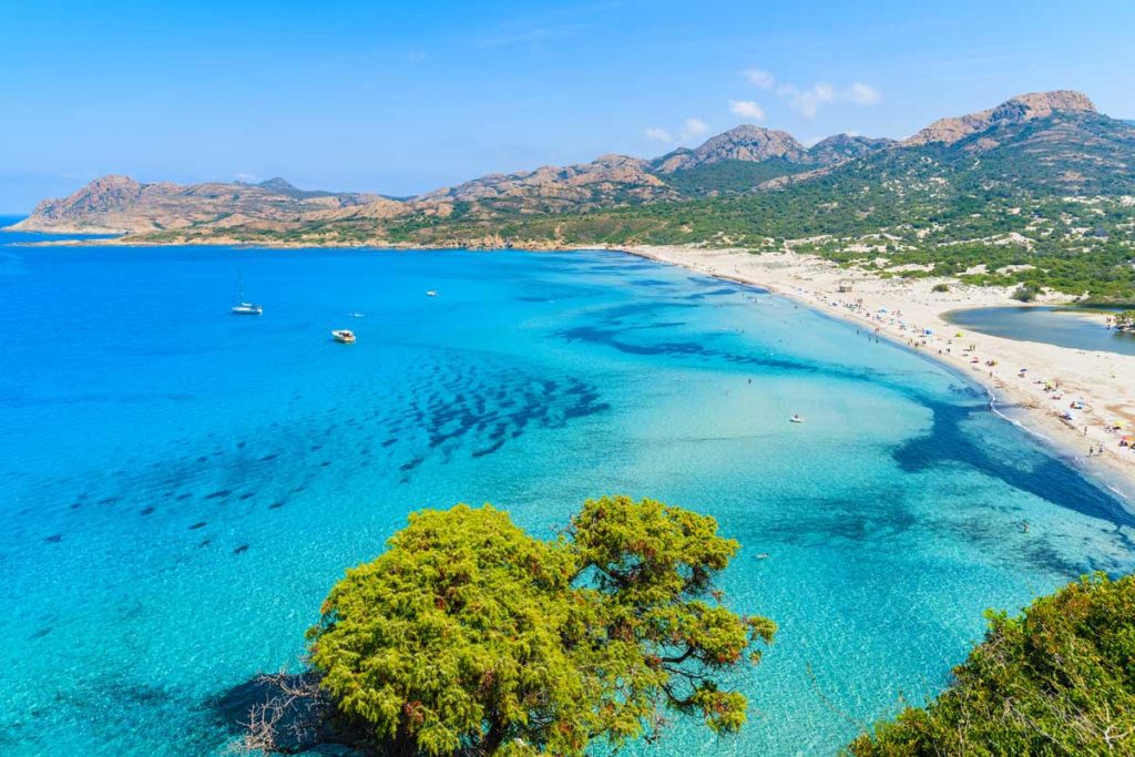 Panoramic view of a pristine beach in the Balagne, Corsica, featuring crystal-clear turquoise water and fine white sand along the Mediterranean coastline.