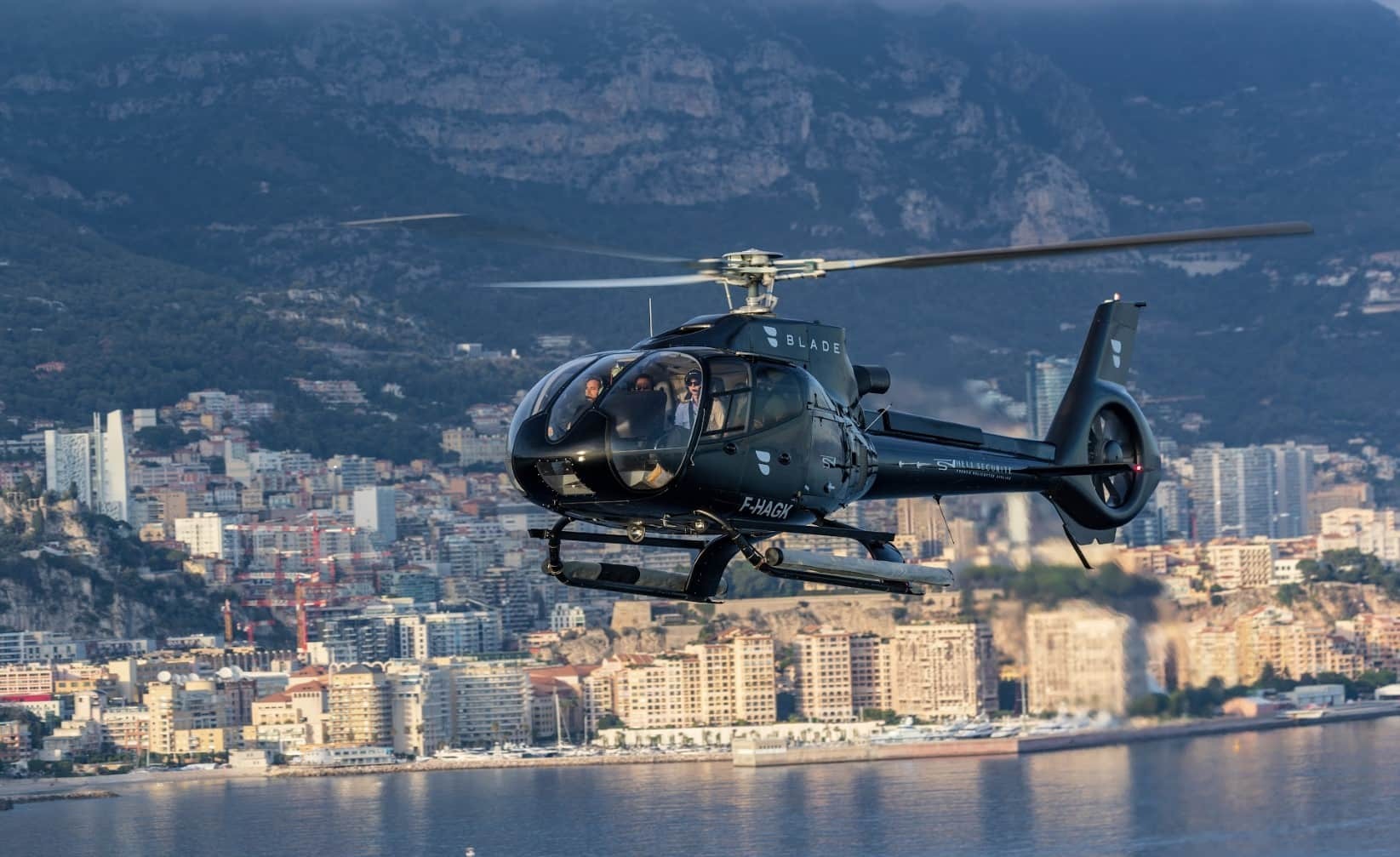 A blade helicopter flying over Monaco’s coastline, with the Mediterranean Sea, and Monte Carlo skyline in the background.